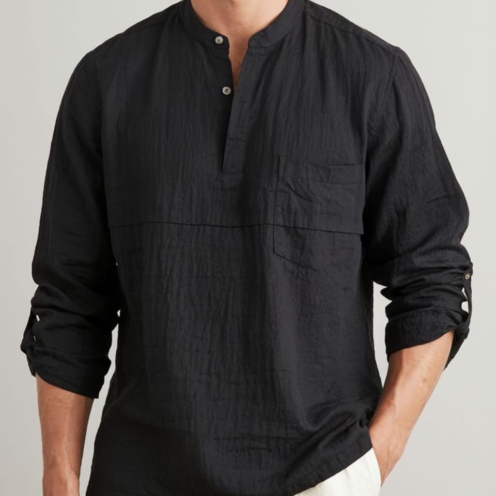 A person wearing a black, long-sleeve, button-up shirt with a stand collar, displayed with sleeves rolled up and their left hand in their pocket. The shirt has a textured, slightly crinkled fabric with a chest pocket visible on the left side.