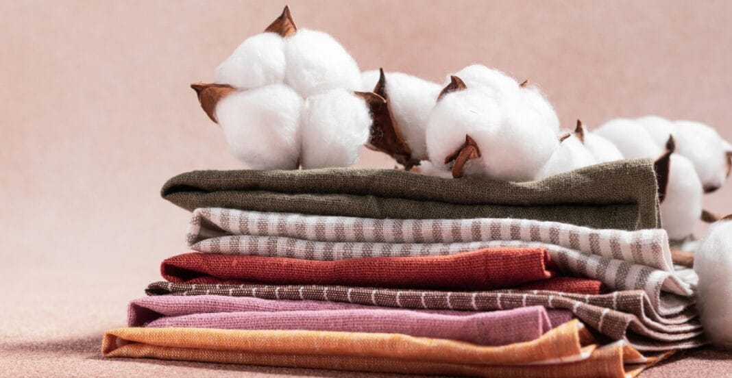A stack of folded cloth napkins in various colors and patterns, placed on a surface with a bunch of cotton plants resting behind them.