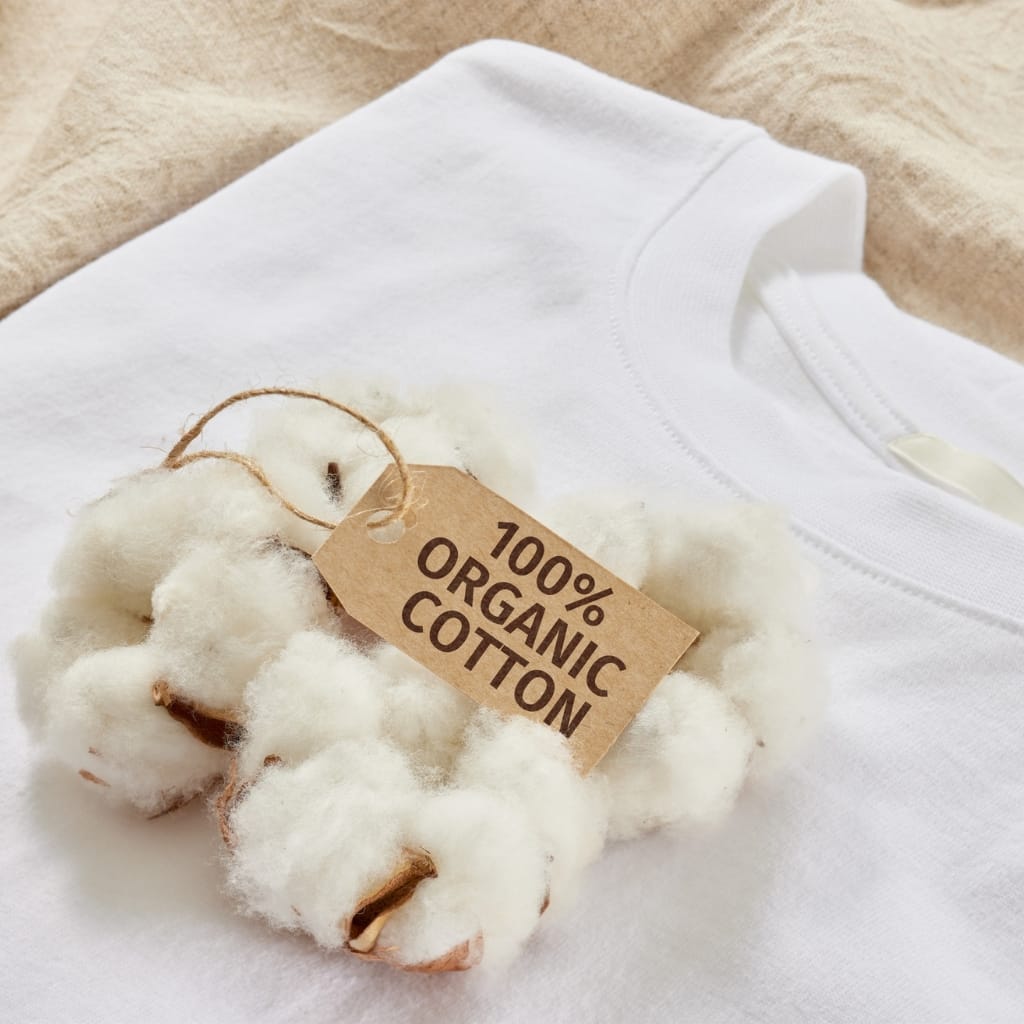 A white cotton T-shirt with a tag that reads "100% Organic Cotton" laid on a light fabric background.
