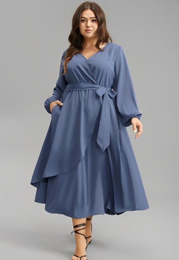 A woman wearing a long, flowing, blue dress with long sleeves, a wrap-style bodice, and a matching belt tied at the waist, designed for a pear-shaped body. The dress features a tiered, full skirt that accentuates the waist and balances wider hips.