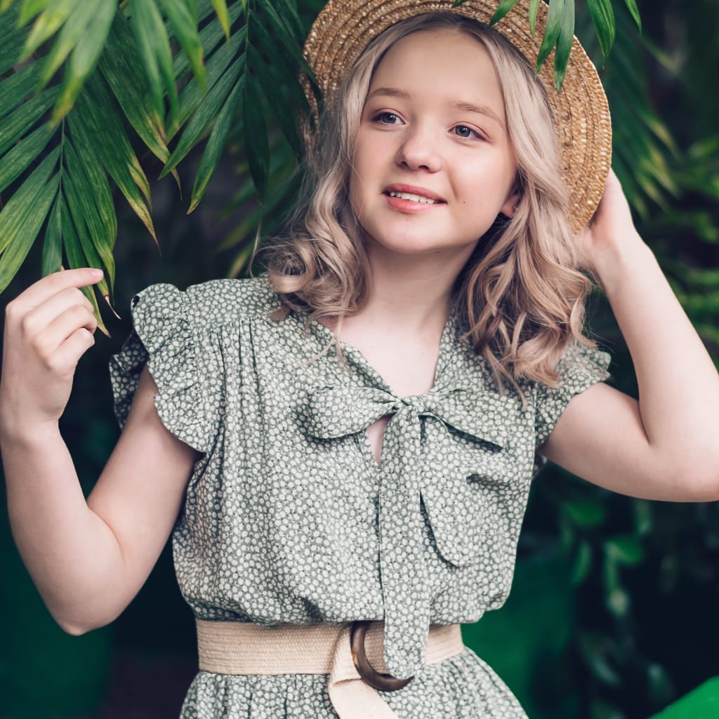 A young girl with blonde, curly hair wearing a green and white patterned dress with puffed sleeves, a bow detail at the chest, a light beige wide belt, and a woven straw hat, standing amidst lush green foliage.