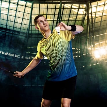 A young male badminton player preparing to serve in an indoor sports arena, wearing a yellow and blue mesh spandex sports shirt.