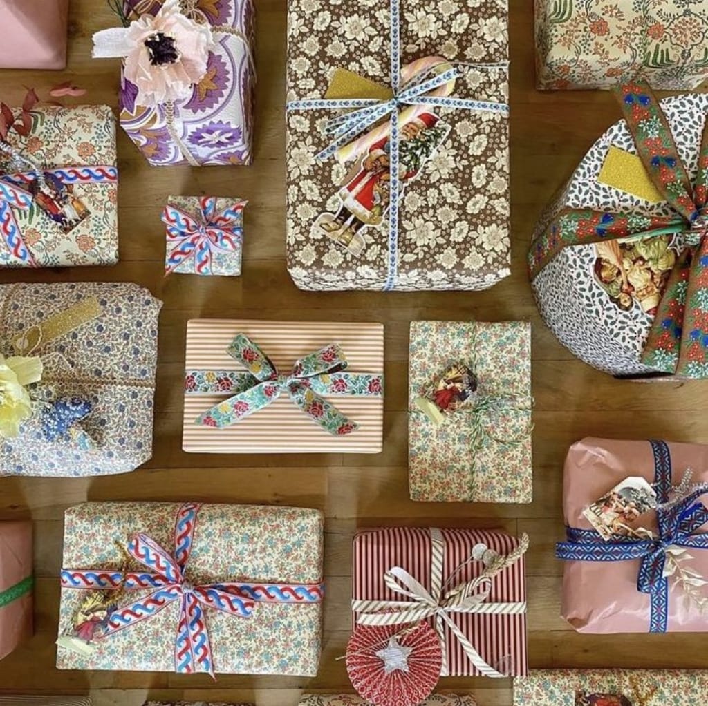 A collection of beautifully wrapped DIY gift boxes and presents, decorated with various colorful cotton print fabrics, bows, and gift tags, arranged on a wooden surface.