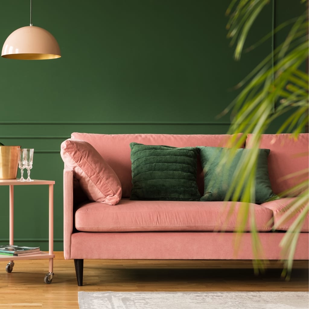 A stylish living room featuring a soft pink velvet sofa with dark green cushions, a pink side table with glasses and a book, a gold bell-shaped pendant light, a green accent wall, and some indoor plants.