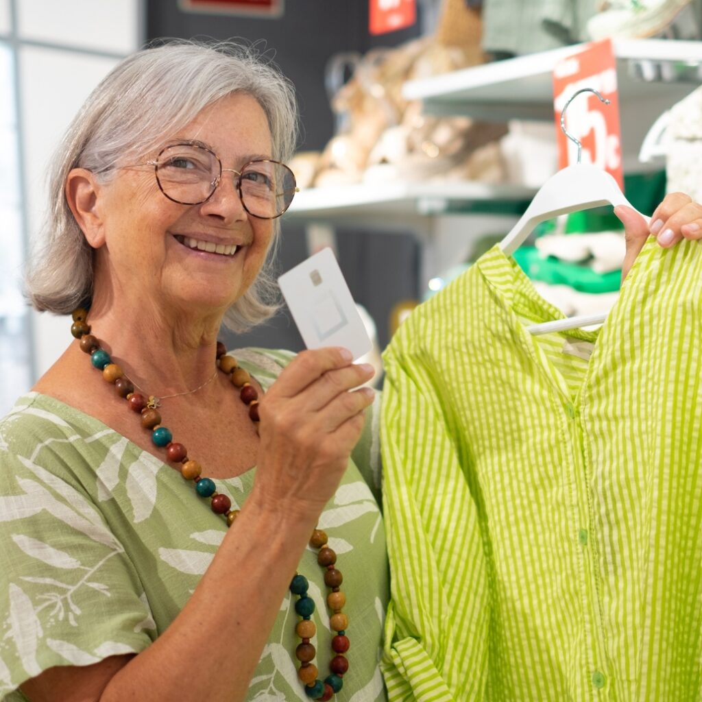 An elderly woman smiling and holding a price tag while shopping in a store, holding a bright green-striped shirt on a hanger.