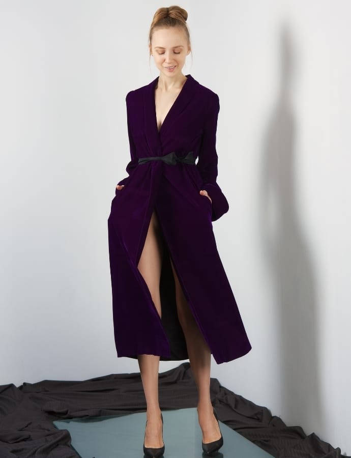 A woman standing against a white wall, wearing a long, deep purple velour coat with a black belt, black high heels, and a subtle smile, with her hands in her pockets.