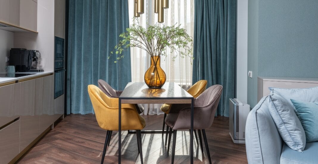 Close-up of a modern home interior featuring a dining table with a large amber glass vase containing a leafy green plant, surrounded by colorful velvet upholstered chairs, with curtains and a cozy sofa in the background.