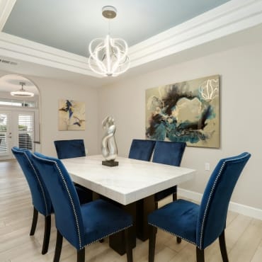 Elegant dining room with a white table, six blue velvet chairs with studded accents, and modern artwork on the walls, illuminated by stylish lighting.