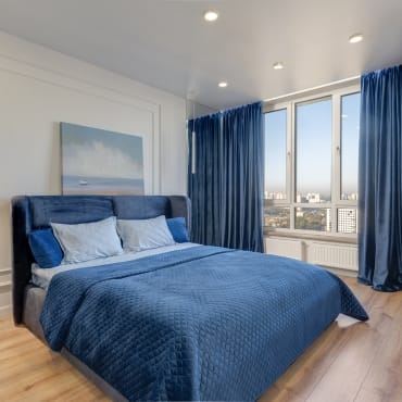 Bedroom featuring a plush blue velvet bedspread, matching velvet pillows, and curtains, with a view of city buildings through large windows.