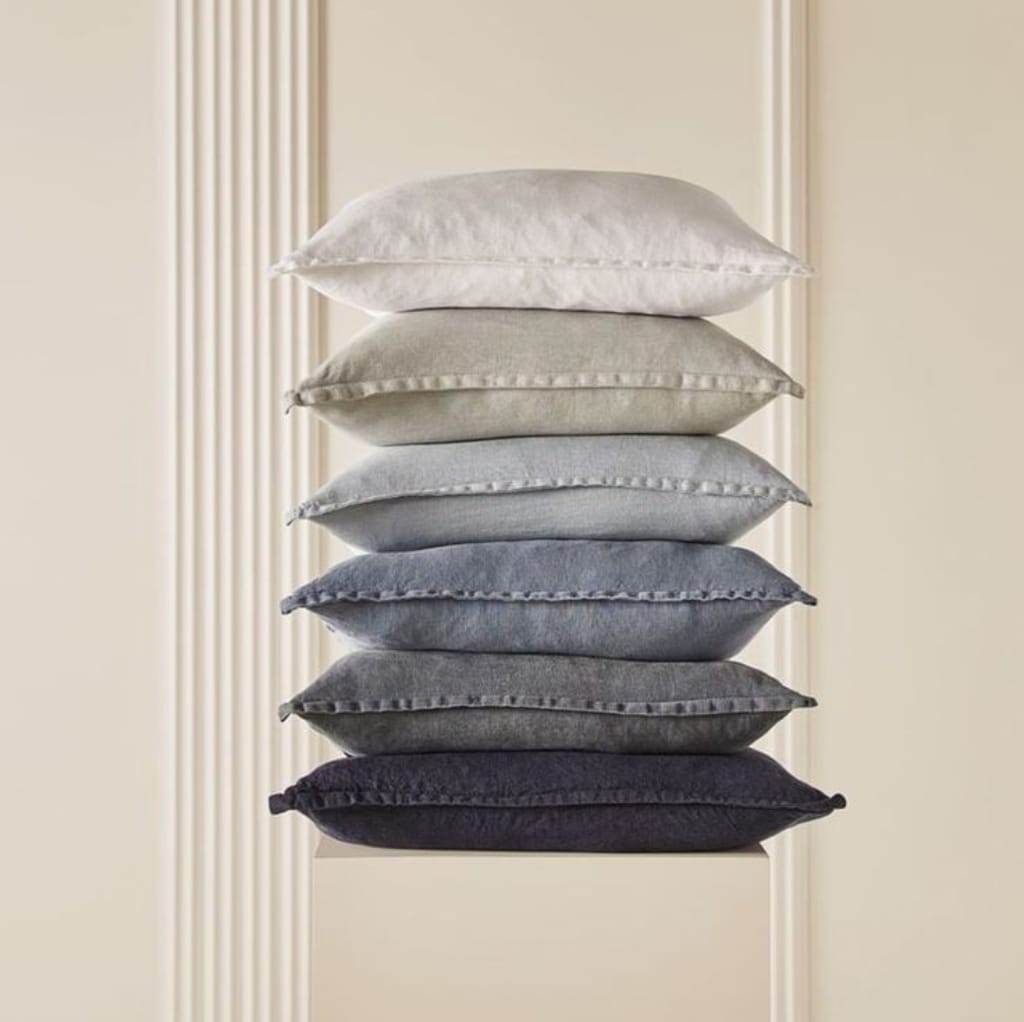 A neatly stacked pyramid of six plush, multi-colored cushions in shades of white, beige, gray, and black, set against a neutral background with vertical wall accents.