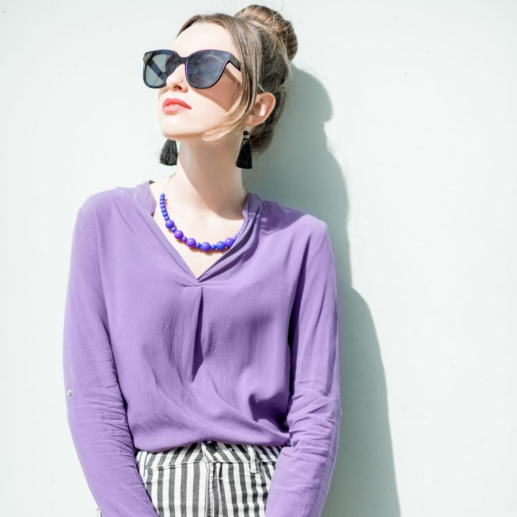 A woman wearing a light purple polyester chiffon blouse, paired with striped pants, accessorized with a blue beaded necklace and black tassel earrings, and sporting large sunglasses against a plain white wall.