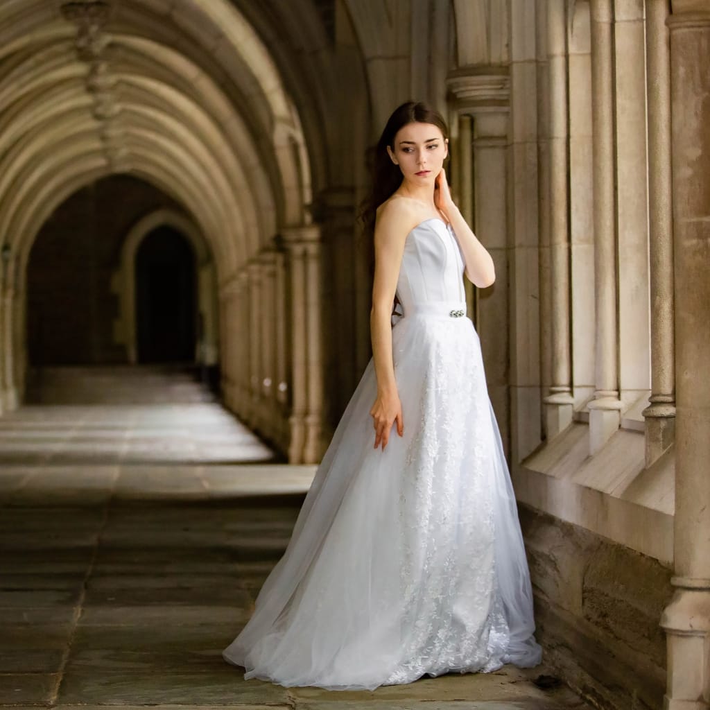 A young woman in an elegant white wedding dress standing in a historic stone corridor with arched architectural details, gazing thoughtfully to her side. The dress has a fitted bodice and a flowing skirt made of polyester organza.