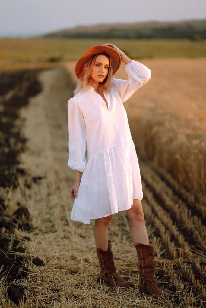 A young woman wearing a white smock cotton dress, cowboy boots, and a wide-brimmed hat standing in a wheat field at sunset.