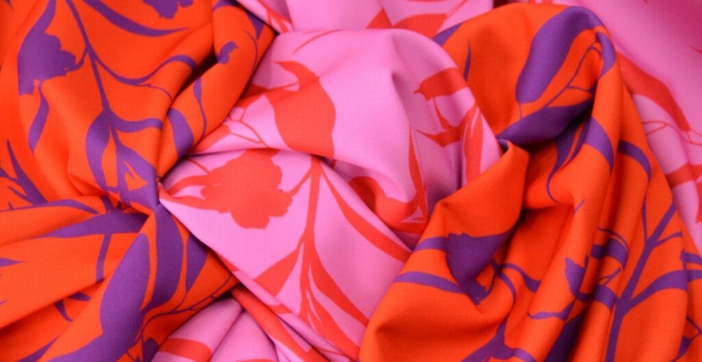 Close-up image of pink and orange chiffon fabric with purple floral or botanical prints, showing the soft, flowing texture and vibrant print design.