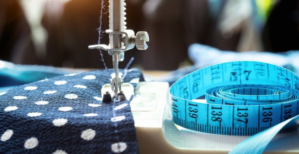 A close-up photo of a sewing machine stitching blue polka dot fabric, with a blue measuring tape coiled on the side.