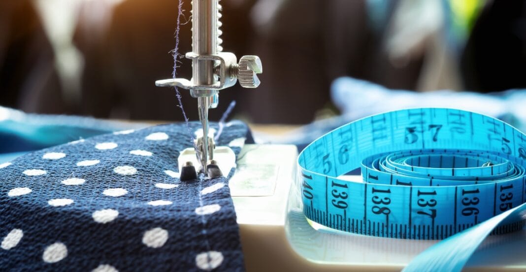 A close-up photo of a sewing machine stitching blue polka dot fabric, with a blue measuring tape coiled on the side.
