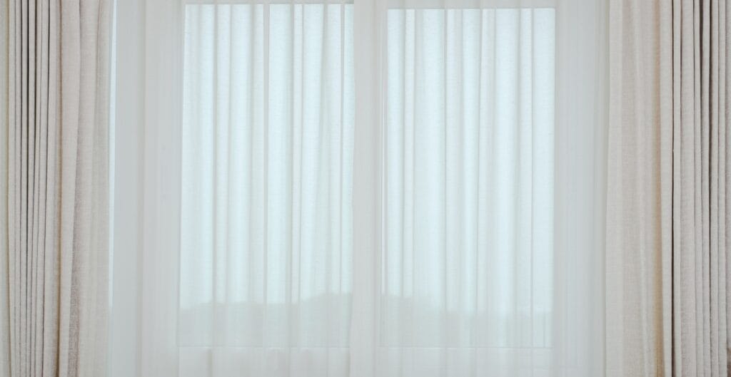 Sheer white curtains with beige drapes on either side, hanging in front of a window.