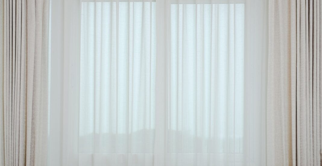 Sheer white curtains with beige drapes on either side, hanging in front of a window.