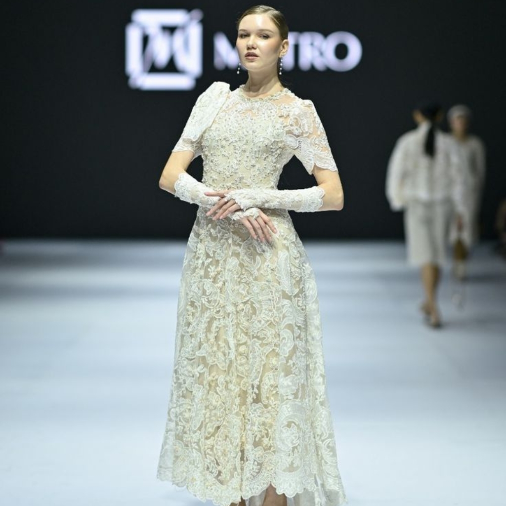 A woman walking on a runway wearing an elegant, intricate cathedral lace dress with matching lace gloves. The background is dark with a logo and some people in the distance.