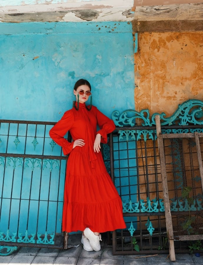A woman in a bright red tiered cotton dress with long sleeves, paired with white sneakers, standing against a vibrant turquoise and weathered orange wall with ornate metal railings. She is wearing sunglasses and statement earrings, with her hand on her hip.