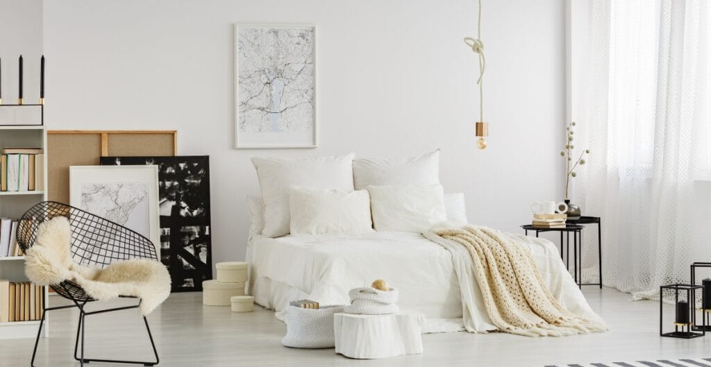 Bright, minimalist bedroom with a white bed, fluffy blanket, black metal side table, bookshelf, framed artwork, and modern décor, all in soft neutral tones.