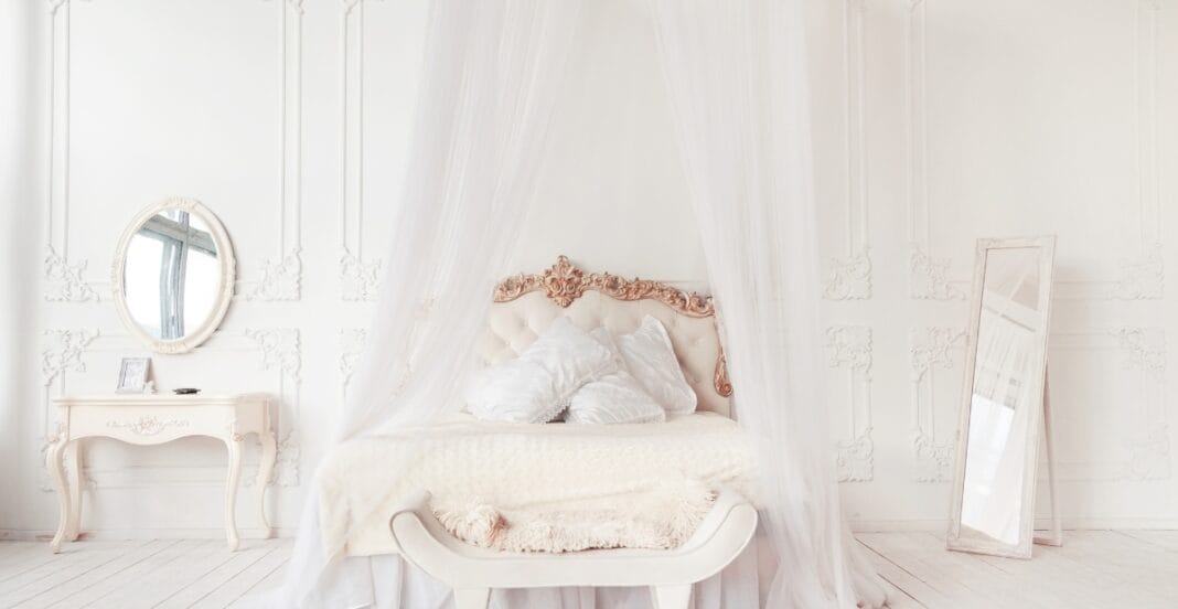 A luxurious bedroom with a white, ornate bed draped with sheer curtains, a matching white vanity table with a mirror, and a full-length mirror leaning against the wall. The room features intricate wall moldings and a soft, elegant atmosphere.