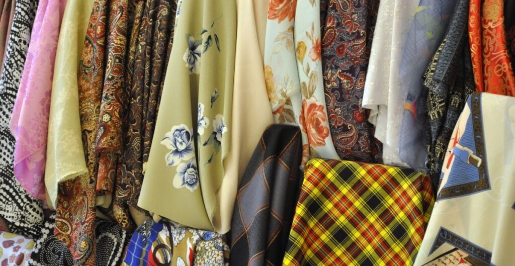 A collection of colorful fabrics hanging side by side, featuring various patterns including floral, paisley, checkered, and abstract designs.