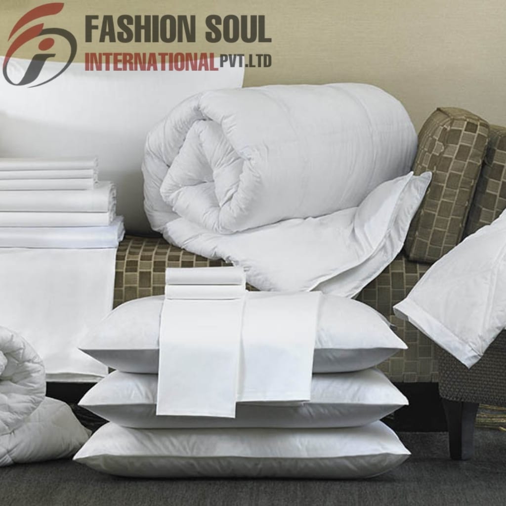 Display of neatly arranged white bed linens, including pillows and rolled-up bedding, stacked on and around a sofa, with a logo and text "Fashion Soul International Pvt. Ltd" in the top left corner.