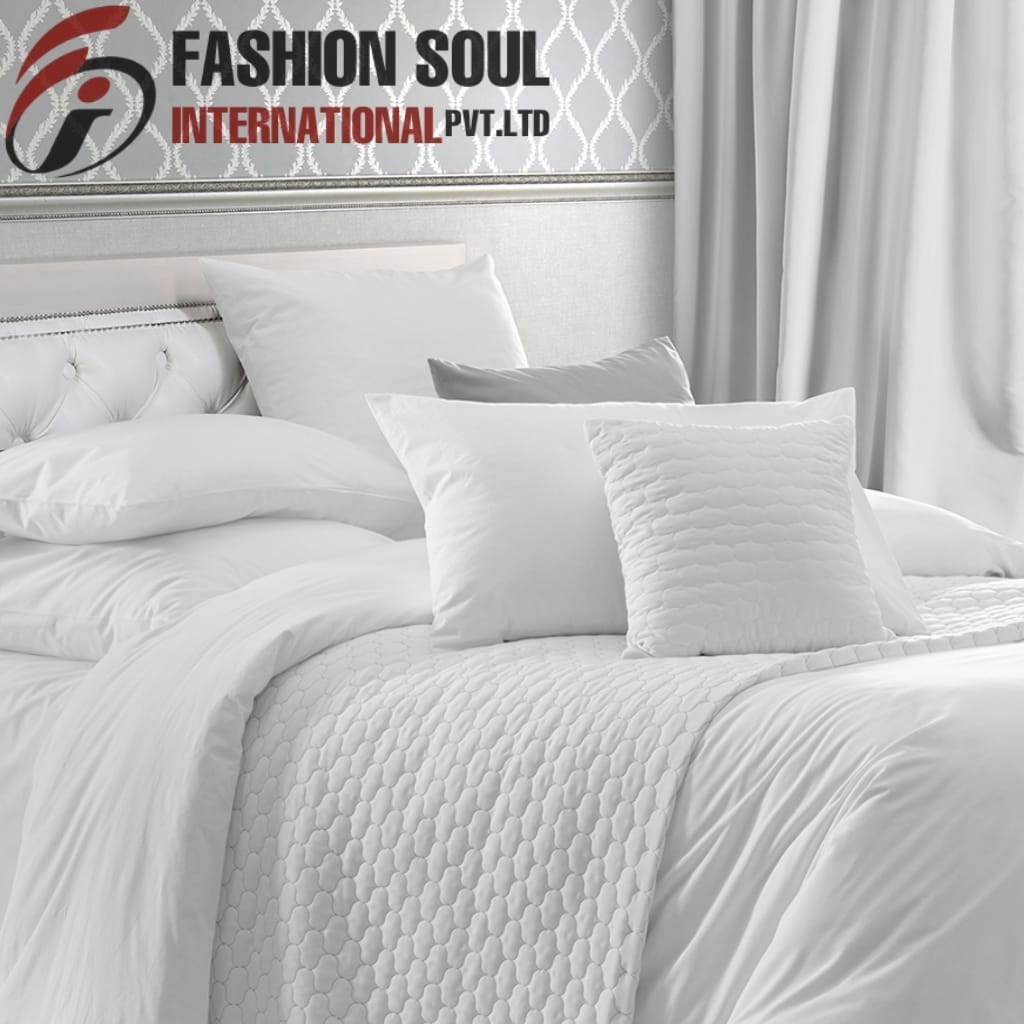 A neatly made white bed with multiple pillows and a textured quilt, set against a wall with patterned wallpaper and curtains. The top left corner features a logo and text reading "FASHION SOUL INTERNATIONAL P.V.T LTD."