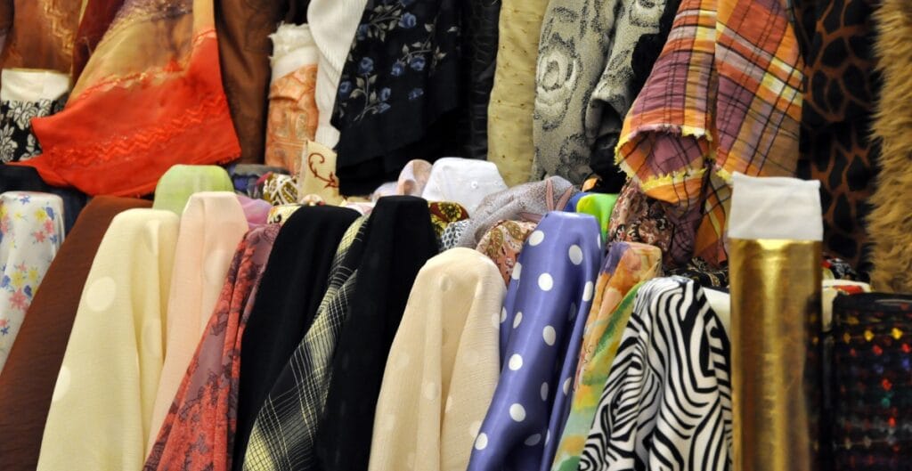 A variety of colorful fabrics and textiles displayed on a market stall, with different patterns including polka dots, stripes, plaid, and floral designs.