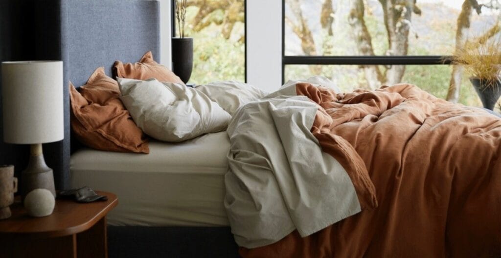 A cozy bedroom with an unmade bed featuring white and rust-colored bedding, a nightstand with a lamp, a book, and decorative objects, and large windows allowing natural light to brighten the room, with trees visible outside.