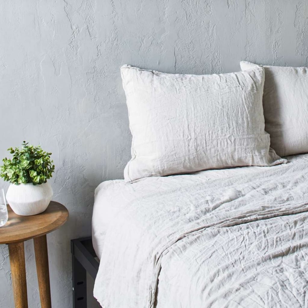 A minimalist bedroom corner featuring a bed with white linen bedding and matching pillow, a textured light gray wall, and a round wooden bedside table with a potted green plant.