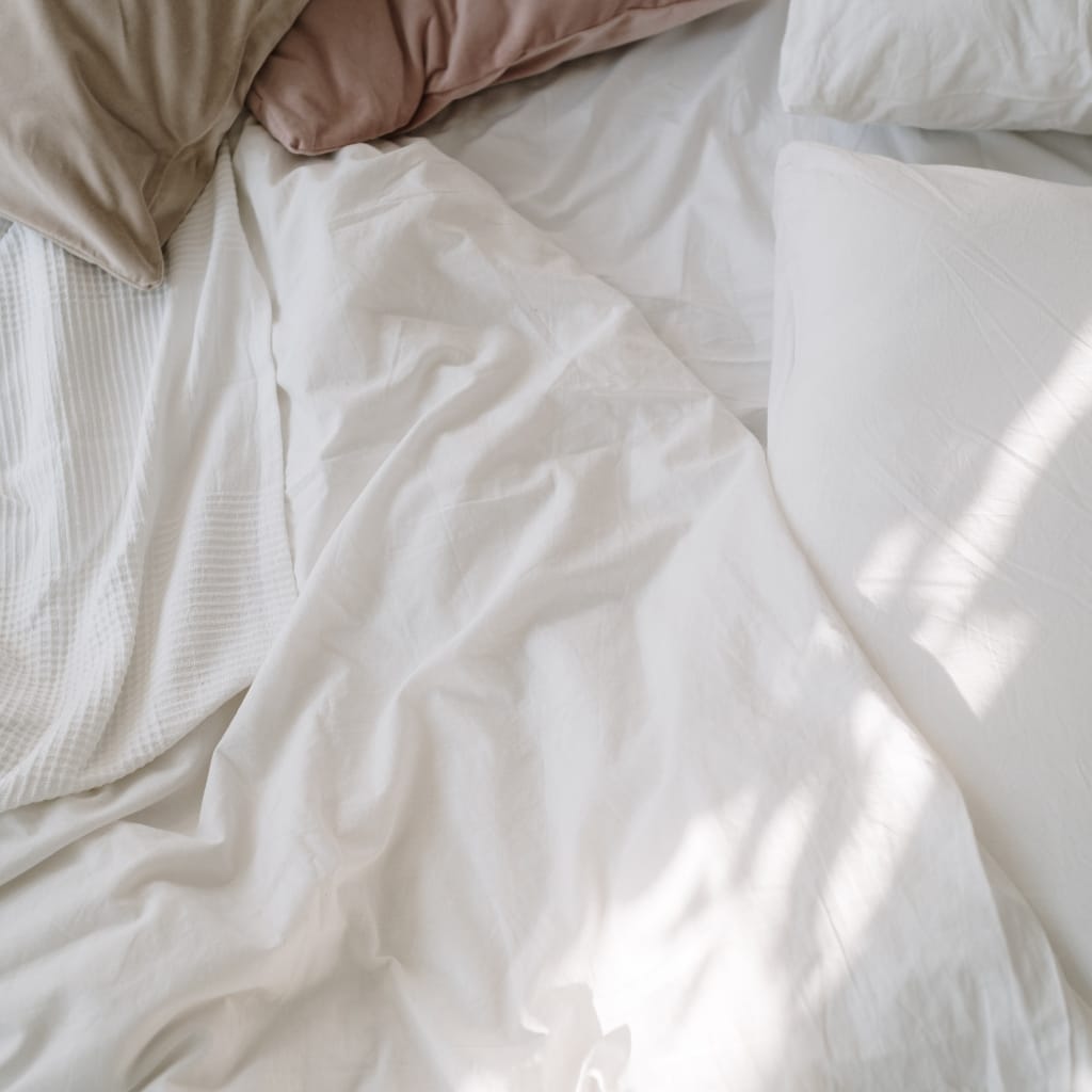 A close-up of unmade beds with white and beige bedding and pillows, displaying a soft, tousled appearance with natural light and shadows.