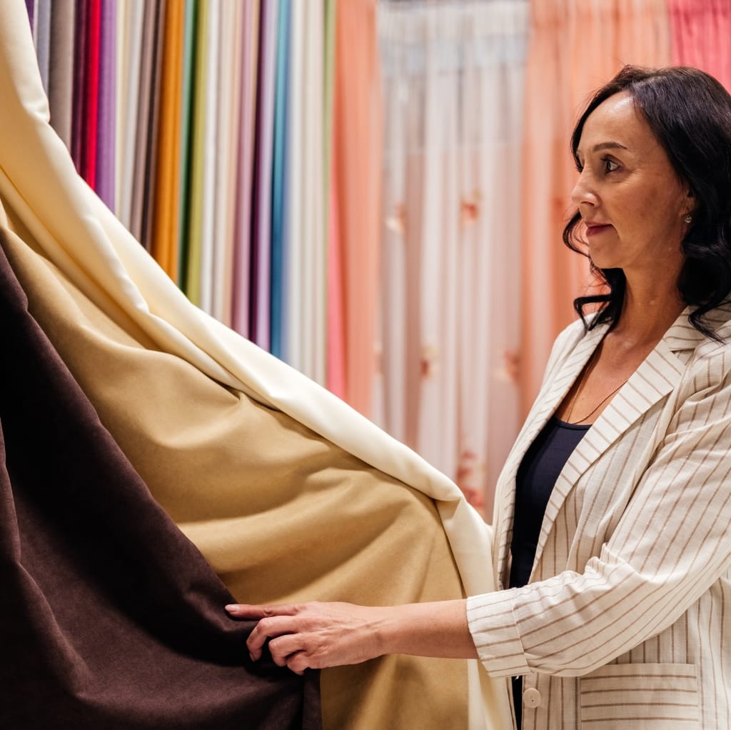 A woman shopping for fabric, holding a piece of beige rayon chiffon against a display of various other fabrics in a fabric store.