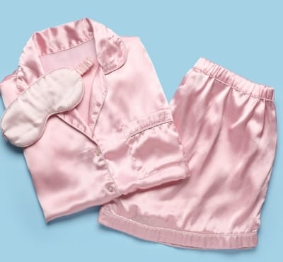 A set of pink satin pajamas including a long-sleeve shirt, shorts, and slippers, laid out on a light blue background.