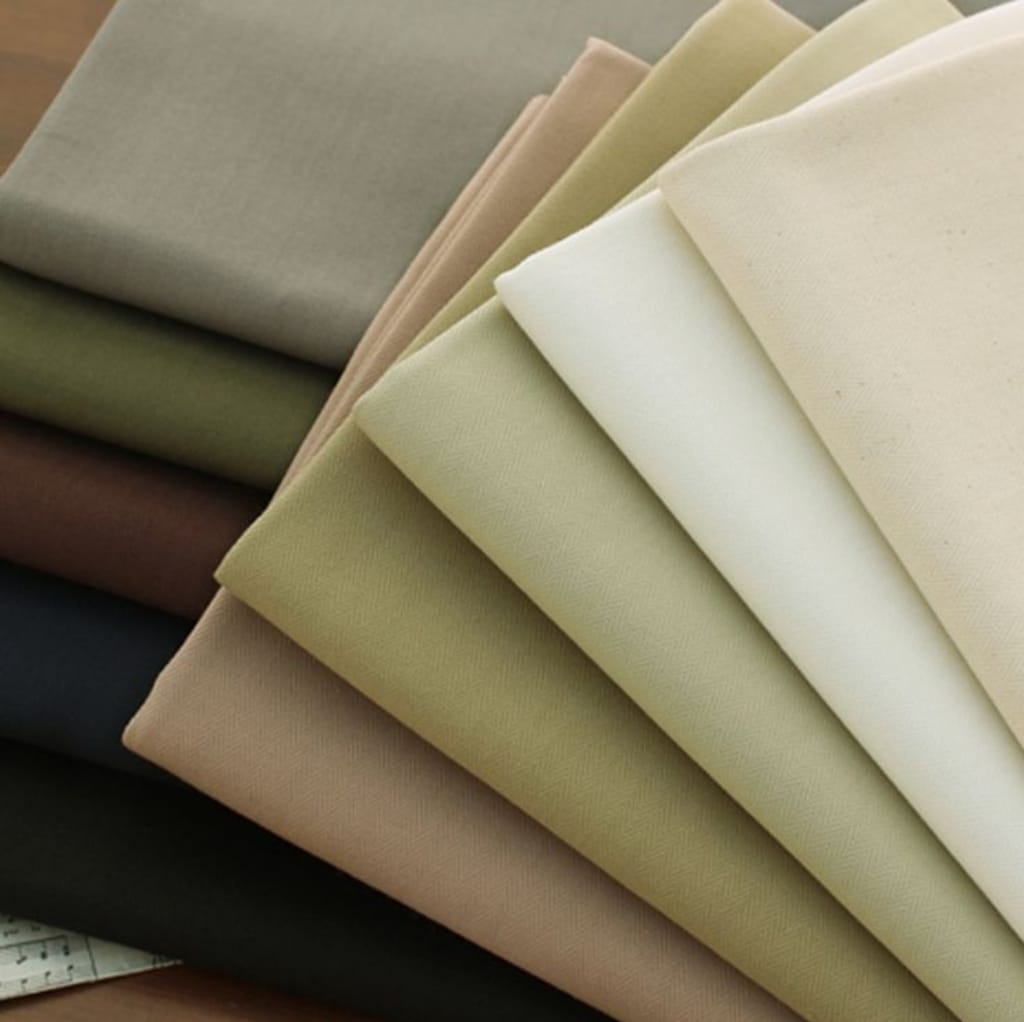 A stack of neatly folded twill-weave cotton fabric in various neutral shades, including beige, light tan, cream, olive green, and dark brown.