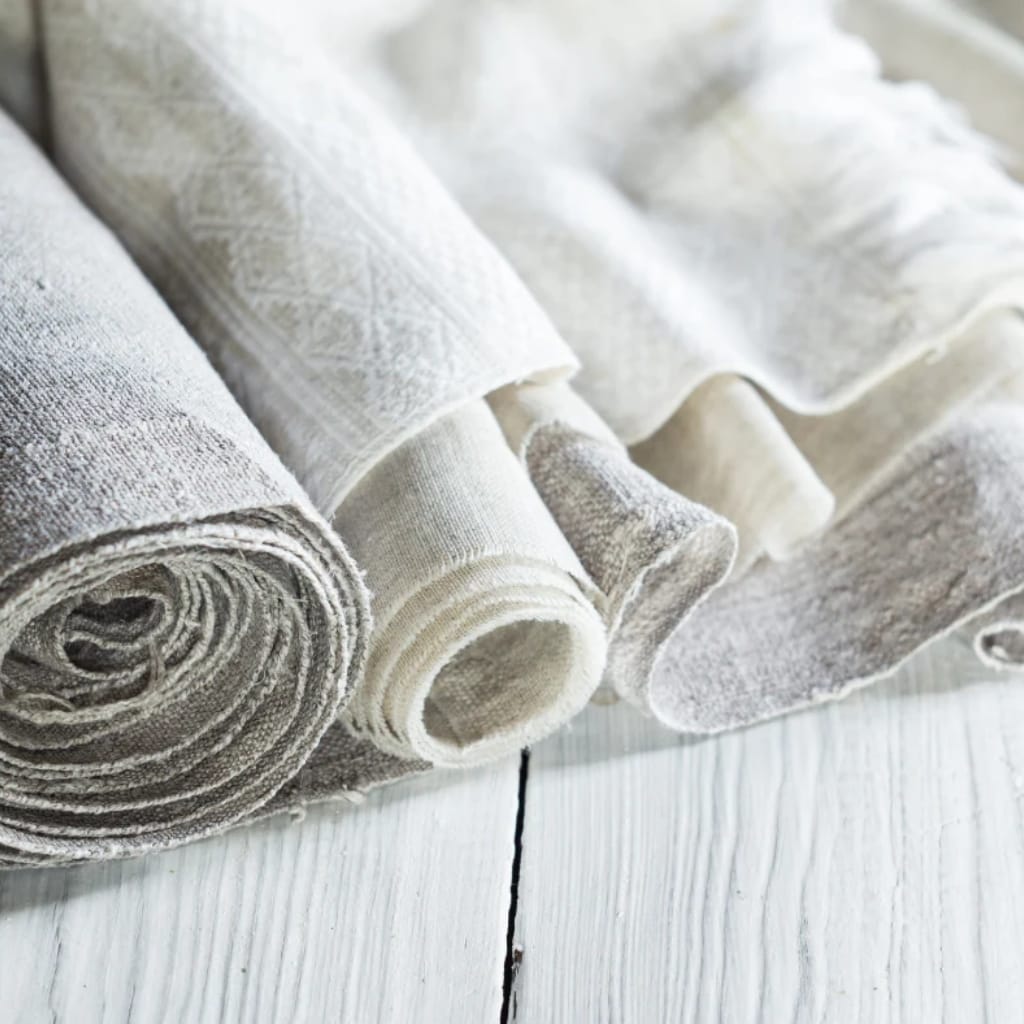Several rolls of light-colored, textured fabric are arranged on a white wooden surface, with some fabric partially unrolled.