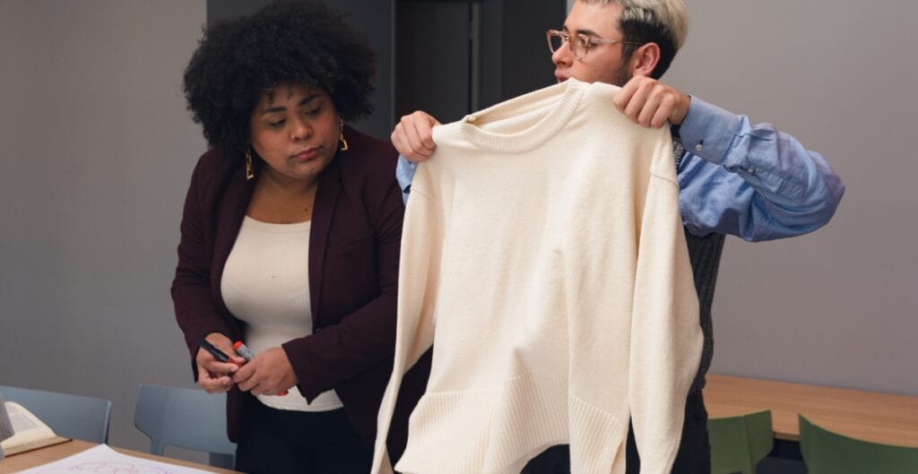 A man and a woman are in an office, with the man holding up a large, cream-colored sweater while the woman looks on attentively, holding a pen.