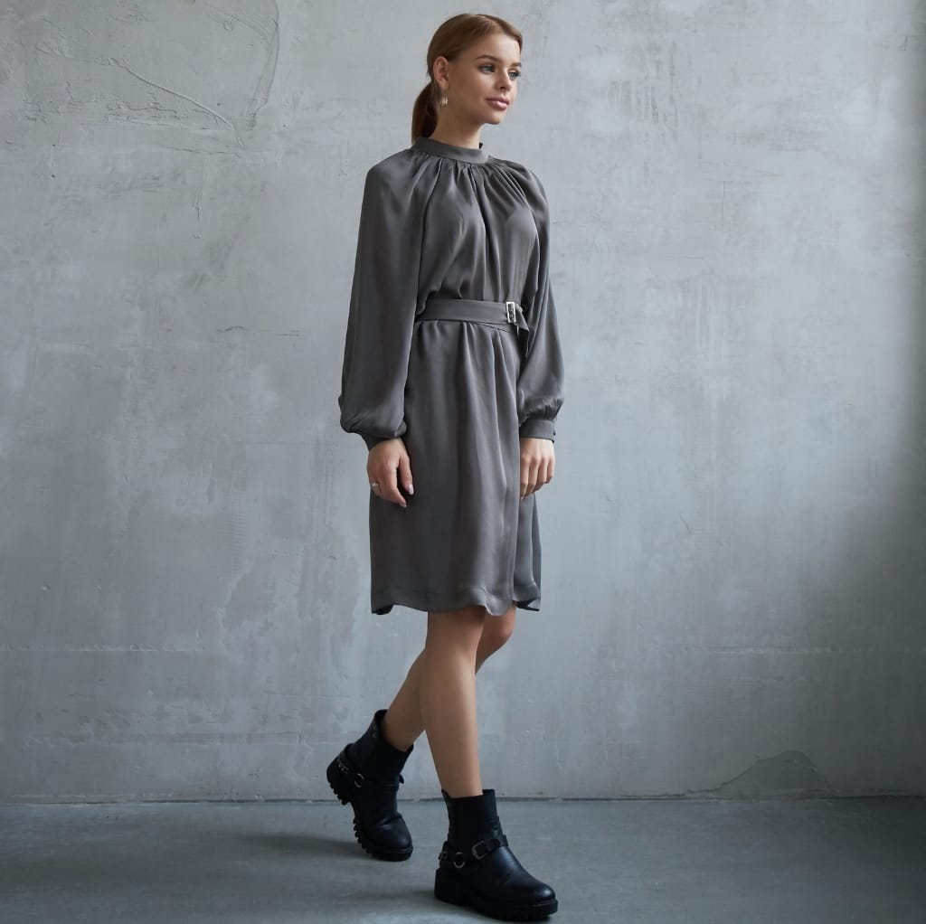 A woman wearing a shiny, gray satin weave cotton dress with long puffed sleeves, belted waist, and black ankle boots, standing against a gray textured wall.