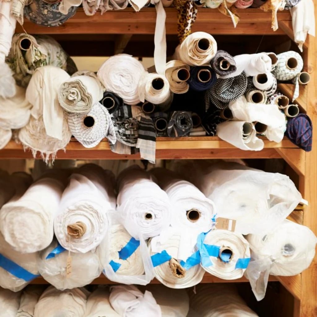 Rolled fabric or textile materials stored on wooden shelves, with various colors and patterns, including white, black, beige, and striped.