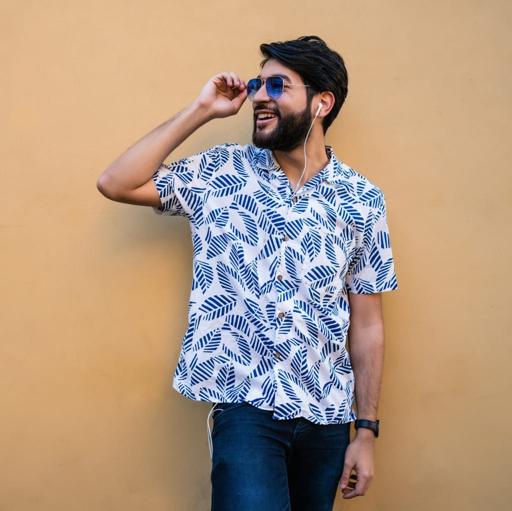 A man with dark hair and a beard, wearing sunglasses, a patterned short-sleeve button-down shirt, and earphones, smiling and leaning against a plain yellow wall.
