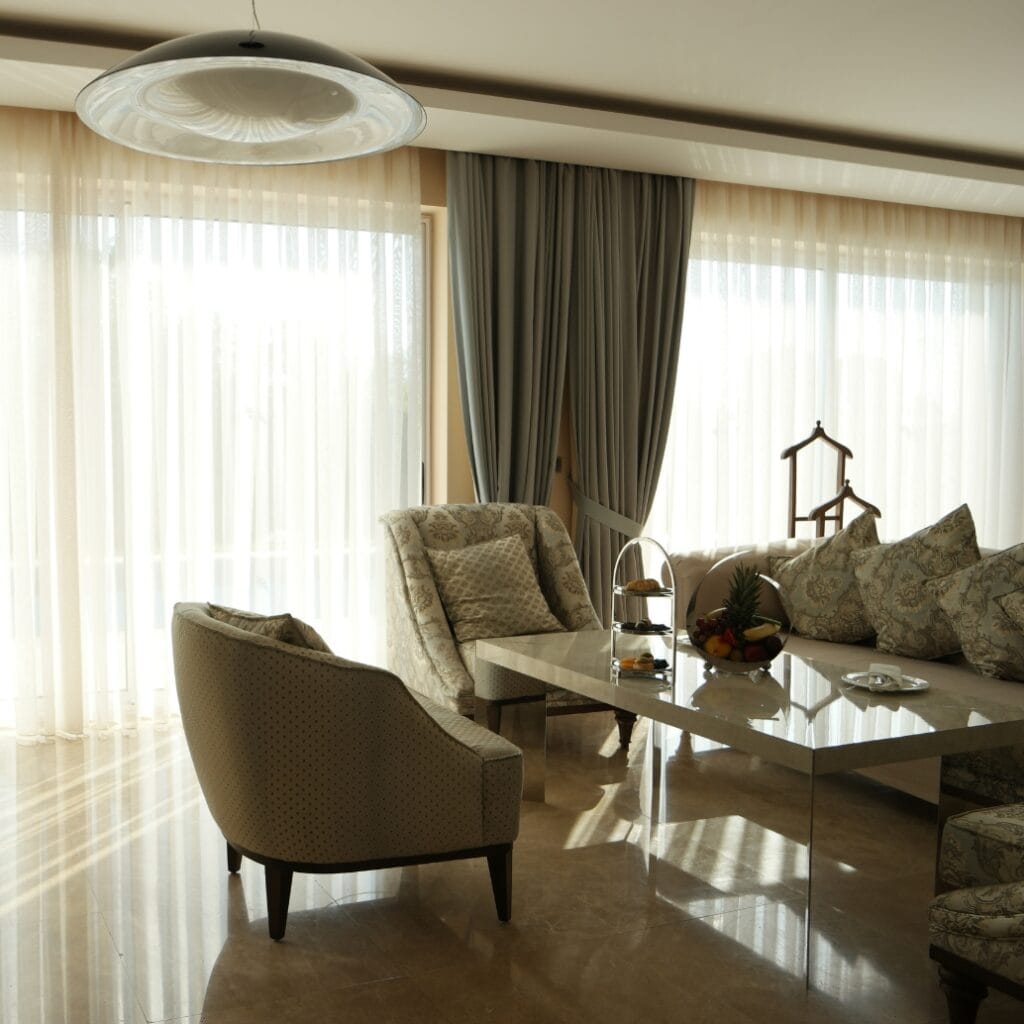 A bright, elegant living room with sheer curtains and floor-to-ceiling windows letting in sunlight. There are upholstered chairs, a marble coffee table with a fruit bowl and snacks, and a sofa with patterned cushions. A ceiling light fixture is visible at the top of the image.
