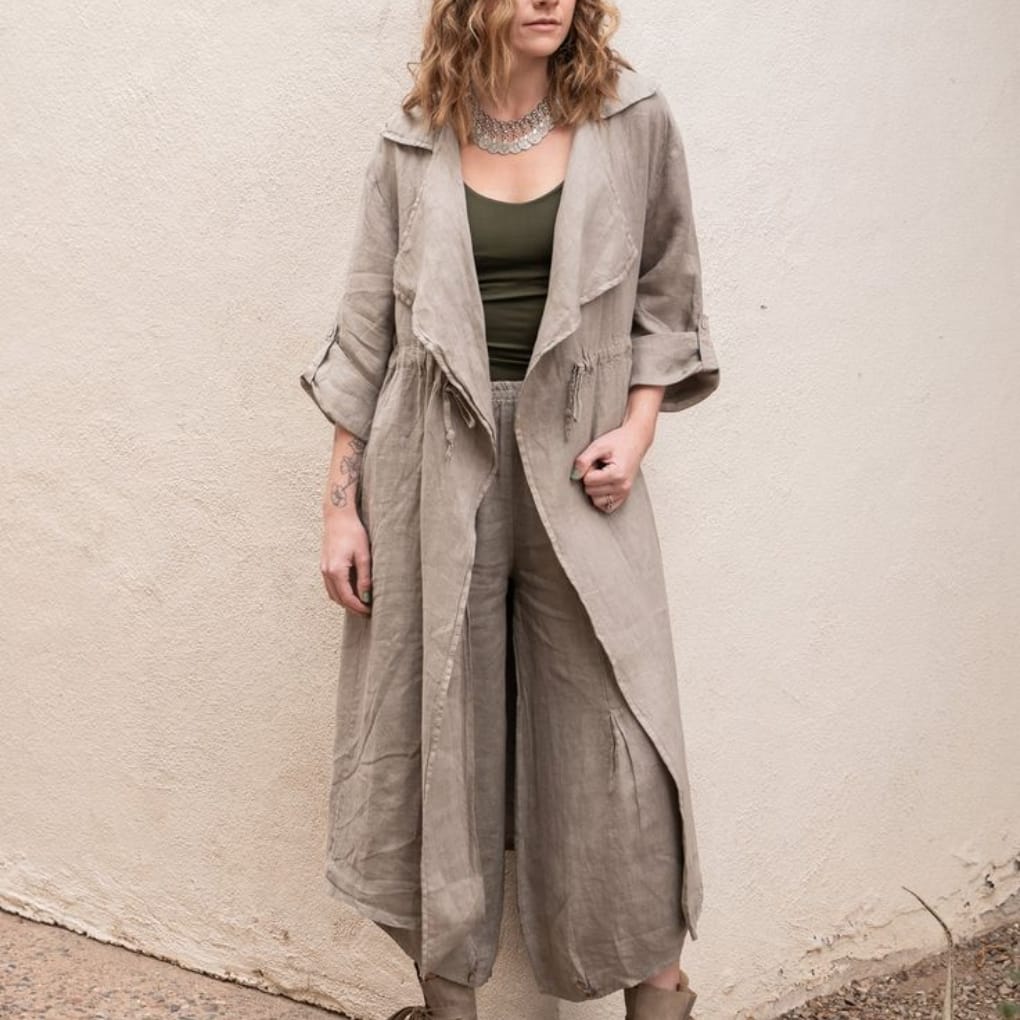 Woman wearing a loose, beige linen trench coat over an olive-green top and pants, standing against a plain neutral wall.