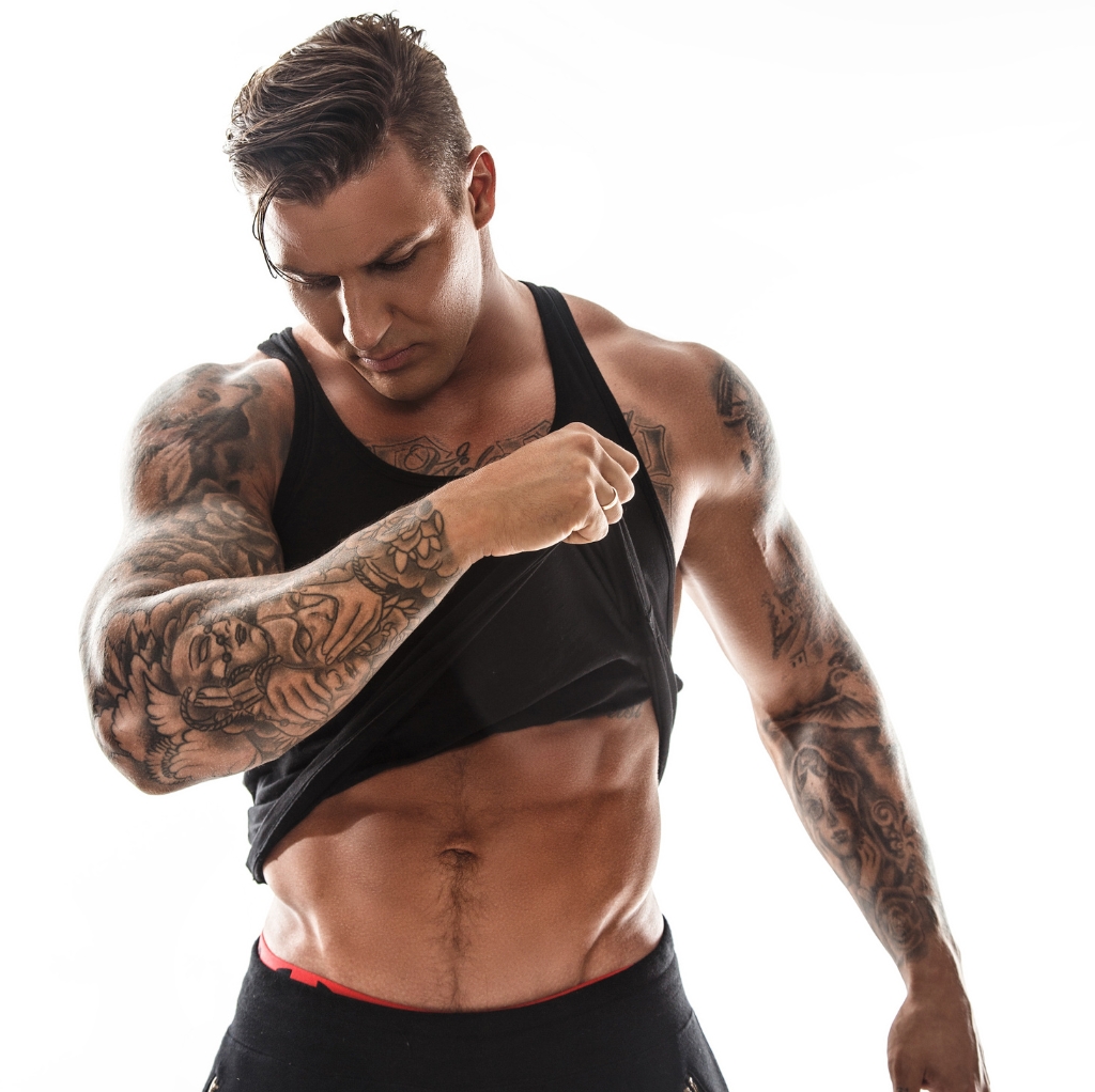 A muscular man with tattoos lifting his black sports tank top to reveal defined abs and chest muscles, standing against a plain white background.