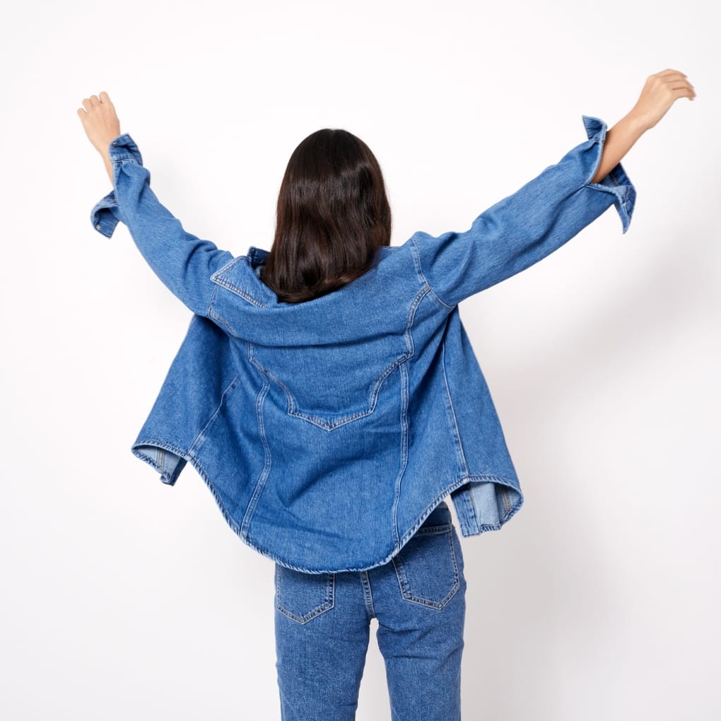 Back view of a woman with shoulder-length dark hair, wearing an oversized denim chambray shirt and matching jeans, standing against a plain white background with arms raised.