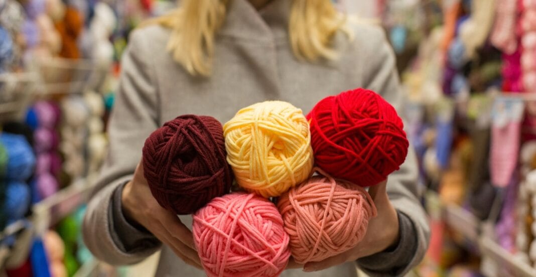 A person holding five balls of yarn in various colors (brown, yellow, red, pink, and peach) inside a craft store.