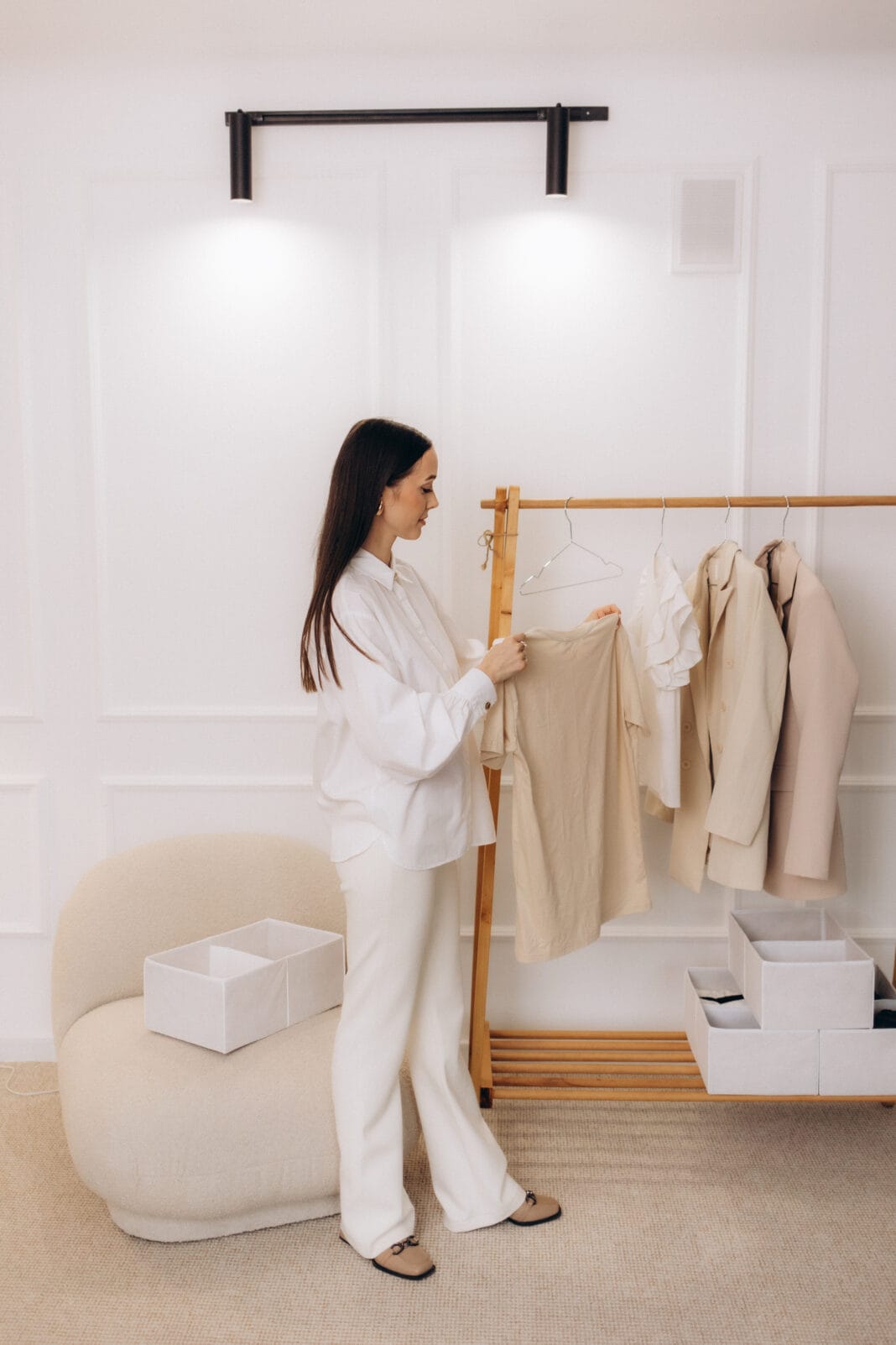 A woman with long dark hair dressed in white pants and a white shirt stands in a minimalist room, looking at and holding a beige piece of clothing on a wooden clothing rack. The room has white walls, a beige carpet, a beige upholstered chair, and white storage boxes.