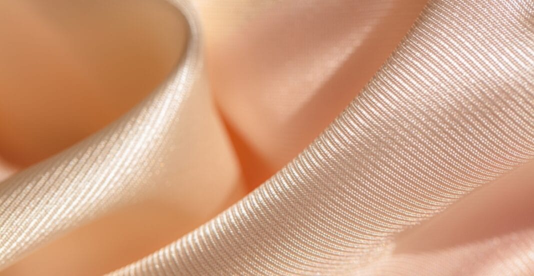 Close-up image of smooth, shiny, peach-colored acetate fabric with a fine, ribbed texture, gently draped to highlight its glossy finish.