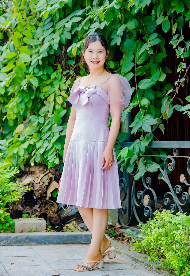 A woman wearing a pastel purple dress with an apple-shaped body silhouette, standing outdoors by a lush green background.
