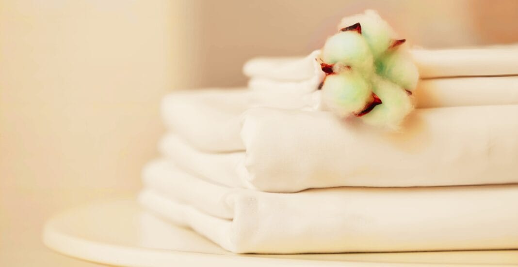 A stack of neatly folded silk sheets with a small cluster of cotton bolls resting on top, against a soft, neutral background.